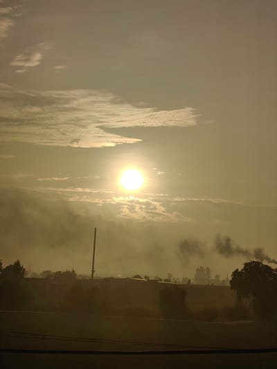 This image depicts a hazy sunset over the industrial landscape of Ganjbar, India. The sun, a bright, hazy orb, is positioned in the upper center of the frame, partially obscured by clouds and atmospheric haze. Streaks of light radiate outwards, illuminating the sky with a warm, golden hue. In the foreground, silhouetted against the fading light, are several industrial structures, including a tall, slender smokestack emitting dark plumes of smoke. The landscape is dotted with trees and what appear to be low-lying buildings. The overall impression is one of industrial activity under a muted, atmospheric sky.