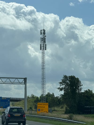 The image shows a cell tower next to a highway in the vicinity of Wierden, Netherlands. The cell tower is tall and metallic, with multiple antennas mounted at various levels. In the foreground, a dark-colored SUV is driving on the highway. To the left of the highway, there are blue directional signs indicating "Almelo-West Wierden-Noord Hardenberg Ommen" and "N36". To the right of the highway, there is a yellow sign that reads "Almelo Centrum Ootmarsum H ZGT vold". The environment is characterized by a cloudy sky and green vegetation, including trees and tall grass. There are also some stacked bales of hay or similar material visible in the background on the right. The scene appears to be captured from a moving vehicle.