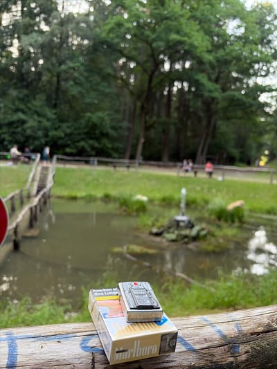 This image, taken in Sohren, Germany, shows a close-up of a Marlboro cigarette pack and a Zippo lighter resting on a weathered wooden surface. The background is softly blurred, revealing a natural setting with lush green trees and a body of water, likely a canal or pond, bordered by grass and a wooden walkway or steps. In the distance, several people are visible, suggesting an outdoor recreational area. The lighting indicates it is daytime, possibly late afternoon or early evening, with diffused light filtering through the trees. The focus is on the objects in the foreground, with the text on the cigarette pack clearly visible, reading "Marlboro" and warning messages in German: "Rauchen schadet Ihrer Lunge" (Smoking harms your lungs). The Zippo lighter also has visible text, including "Zippo 22" and "BRADFORD, PA MADE IN USA". The overall impression is a still life composition set against a tranquil, outdoor backdrop.