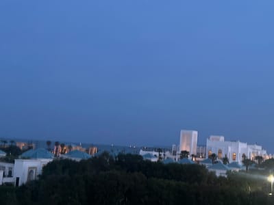 The image shows a twilight view of a coastal resort or residential area in Fnideq, Morocco. The sky is a dark blue, indicating either late evening or early morning.
In the foreground, there is a line of dark green trees partially obscuring the view of the buildings.
Behind the trees are several white buildings with distinctive architecture, possibly in a Moroccan or North African style. These buildings have blue domes and are likely part of a resort or hotel complex. Some of the buildings appear to have illuminated windows and archways.
In the background, there is a body of water, likely the Mediterranean Sea, with faint lights visible in the distance, possibly from ships or coastal cities. Palm trees are interspersed among the buildings and along the coastline. A street light is visible to the right.