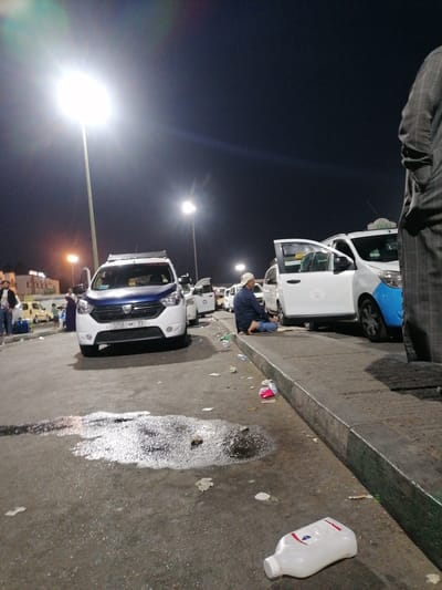 In the city of Inezgane, Morocco, at night, a street scene unfolds. A white Dacia Lodgy taxi with a blue hood and roof rack is parked prominently in the foreground, its license plate reading "3258 | 33". The vehicle's front doors are open. To the right, another white taxi, possibly a Dacia, is also present, with its driver's side door open. A man wearing a light-colored cap and blue clothing is seated on the ground next to this taxi, appearing to be resting or engaged in prayer.
Further back, a group of people are visible, some standing and others possibly queuing near other vehicles. A tall streetlamp illuminates the area, casting a bright light that creates lens flares. The ground is asphalt, with some puddles reflecting the light, and scattered litter is visible. In the far right of the image, a person in a checkered suit stands, partially obscuring the view of the taxis. A white plastic bottle lies on the ground in the foreground, near a discarded cigarette butt. The overall environment suggests a taxi rank or a similar transportation hub at night.