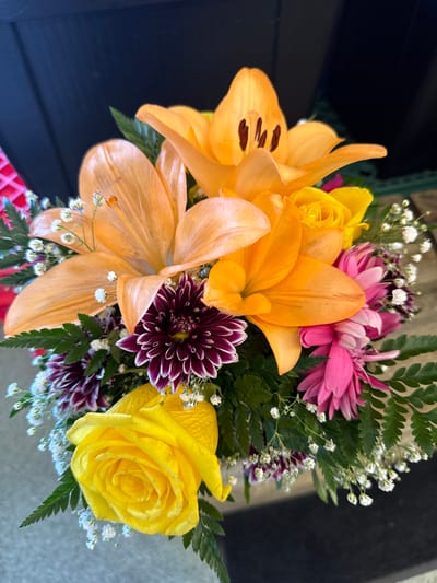 This is a medium shot, overhead, eye-level, close-up, and high-angle medium shot photograph of a bouquet of flowers. The subjects of the photo are the flowers themselves, which are arranged in a cluster, filling most of the frame. The flowers are of various types and colors, including large, bright orange lilies with prominent stamens, yellow roses with layered petals, pink chrysanthemums, and deep purple dahlias with white tips. There are also small white filler flowers, likely baby's breath, and green fern fronds interspersed throughout the bouquet. The lighting suggests it may be daytime, possibly outdoors given the natural light. There are no people or animals visible. The background is dark and out of focus, making the vibrant colors of the flowers stand out. To the left, the dark background appears to be a dark surface, possibly a table or a wall. To the right, the background transitions to a lighter, textured surface, which could be a sidewalk or a concrete patio. A hint of a red, woven pattern is visible at the bottom edge of the frame, possibly part of the vase or container holding the flowers. No text is visible in the image. The overall impression is a colorful and fresh floral arrangement, likely a gift or a decoration.