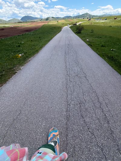 A first-person low-angle perspective captures a person on a skateboard moving along a paved road in Javorje, Montenegro, under a partly cloudy sky. In the immediate foreground, the lower portion of a person's body is visible, wearing a multi-colored plaid shirt (pink, blue, green) and dark green pants. Their left foot, shod in a dark sneaker with white laces, rests on a skateboard featuring a vibrant abstract design in blues, oranges, yellows, and black.

The asphalt road, dark gray and textured, extends straight into the distant landscape. It exhibits significant longitudinal cracking, particularly on the right side. Green grassy verges flank both sides of the road, with small clusters of yellow flowers visible on the left verge.

Beyond the road, the rural environment unfolds under a bright blue sky dotted with scattered white cumulus clouds. On the left, a large expanse of tilled, reddish-brown earth contrasts with wide green fields. In the mid-distance on the road, a white car is visible driving away from the viewer, while a dark farm vehicle, possibly a tractor, is parked off the right side of the road. Further into the background, rolling green hills and mountains rise, some appearing forested. Numerous small, distinctively shaped structures, characteristic of traditional "katun" huts, are scattered across the distant green hillsides on the right. Sparse bushes and trees are also present in the landscape.