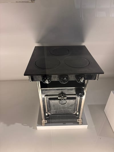 This media file shows a close-up, tilted shot of a vintage black stove with a metal frame, positioned against a plain, light-colored wall. The stove has a flat top with three circular heating elements, and a small oven door is visible on the right side. Three circular control knobs with white indicators are mounted on the front panel of the stove, below the heating elements. These knobs appear to be set to different levels, indicated by numbers ranging from 0 to 4.
The lighting in the image is diffused, casting soft shadows on the wall, suggesting an indoor setting, possibly a museum or an exhibition space, given the clean presentation. The overall tone is neutral, focusing on the object itself. The environment appears to be dimly lit, which highlights the texture of the wall and the details of the stove.
There is text visible on a vertical metal strip on the far left of the image, which reads "Boos + Hohn, Ortenberg, Baden". This text provides a manufacturer or origin cue. The image orientation is slightly tilted, giving a dynamic perspective of the stove. The scene does not depict any people or specific activities, but rather presents a static display of a historical kitchen appliance. The location context is Bern, Switzerland, but this information is not visually evident from the image itself.