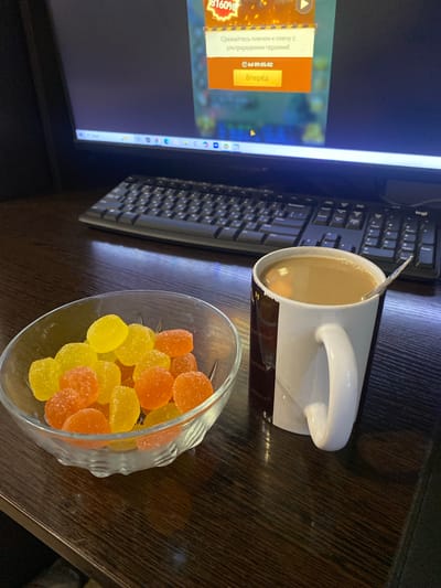 The image shows a desk with a computer monitor, keyboard, a mug of coffee, and a bowl of gummy candies. The monitor displays an advertisement for a game with Russian text, including "Сражайтесь плечом к плечу с ультраредкими героями!" (Fight shoulder to shoulder with ultra-rare heroes!) and a button that says "Вперёд" (Forward). A clock icon shows 09:05:02. The keyboard is a black QWERTY layout. The mug is white with a brown logo. The gummy candies are orange and yellow, and appear to be a type of jelly candy. The setting appears to be indoors, likely a home office or study, given the presence of a computer and casual items like candy. The lighting suggests it might be morning, aligning with the time displayed on the screen. The overall mood is relaxed and perhaps indicative of a break or starting the day. The wooden desk surface is visible. The location context is Рубцово, Russia.