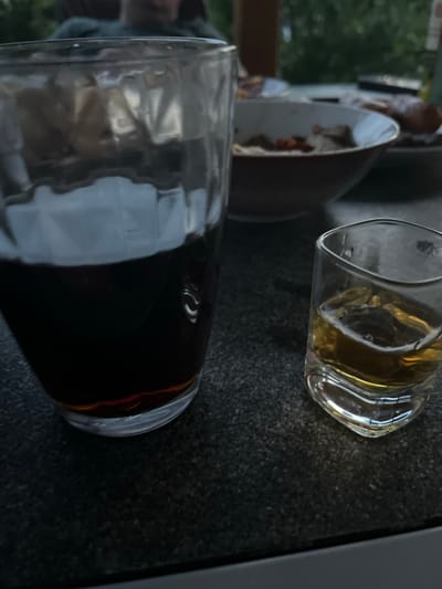 The image shows two glasses of drinks on a dark, speckled surface, possibly a countertop or table. The glass in the foreground is larger and filled with a dark liquid, possibly cola or a dark spirit, with ice cubes visible. The glass in the background is smaller and contains a lighter, amber-colored liquid, likely whiskey or another spirit, also with ice. The reflections in the glass in the foreground suggest it is near a window or open space, with greenery and possibly a building visible in the blurry background. The lighting suggests it could be daytime, possibly late afternoon or evening due to the dimmer natural light. The overall scene evokes a relaxed, informal setting, perhaps a home or a casual bar. There is no visible text in the image. The location context provided is Milná, Czechia.