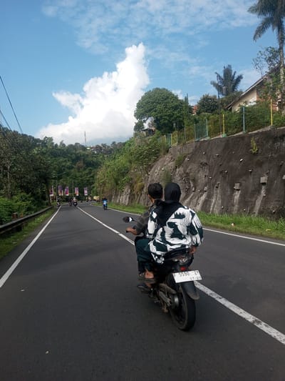 A motor scooter carrying two people travels along an asphalt road. The passenger in the back wears a black head covering and a white and black patterned garment. The license plate reads "F 5590 ZY 07.23". The road has a solid white line marking its edge, and a broken white line separating lanes. On the left of the road is a metal barrier. Across the road is a slope with a concrete retaining wall. The sky is blue with large white clouds. The location cannot be identified as Khe Luang, Laos.