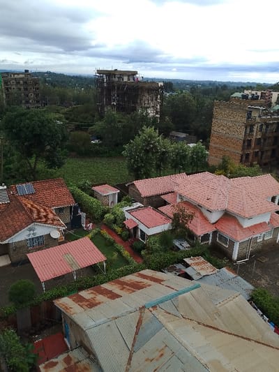 The image depicts an elevated view of a residential and mixed-use area in Olkeri ward, Kenya, under an overcast sky. In the immediate foreground, corrugated metal rooftops, some with visible rust, slant downwards from the bottom edge.

The midground features a cluster of residential structures, predominantly single-family homes or smaller multi-unit dwellings, characterized by red tiled roofs and white or stone/brick facades. Several properties exhibit manicured green lawns, shrubbery, and paved pathways. One house features solar panels on its red roof and a satellite dish on an exterior wall. Small outbuildings with red roofs are visible within some properties. Adjacent to these residences is a large, irregularly shaped green agricultural field or undeveloped plot.

In the background, multiple multi-story buildings are visible. To the left and center-left, two prominent structures appear to be either under construction or in a state of disrepair, featuring exposed concrete frames and scaffolding. To the right, another multi-story building with a reddish-brown brick facade stands. Beyond these structures, a distant horizon reveals further buildings and an expanse of green landscape under the cloudy sky. A white vehicle is partially discernible through trees in the mid-background. No individuals or specific actions are discernible within the frame.
