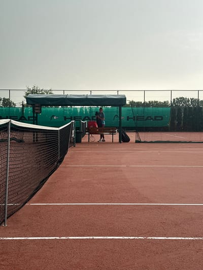 An outdoor tennis court is depicted in Giebułtów, Poland, under an overcast sky with visible light precipitation. The court surface is red with regulation white lines, appearing damp. A black tennis net occupies the left foreground. On the far side of the court, approximately centered horizontally, a single individual stands beneath a green-canopied shelter. The person is dressed in athletic attire, including a dark top and light shorts, and is holding a dark fabric object, potentially a towel.
Adjacent to the individual under the shelter, a light-brown wooden bench is present. A red sports bag rests on the bench to the individual's left. A dark-colored tennis racket lies on the court surface directly in front of the bench. A black sports bag is positioned on the ground to the individual's right. A single yellow tennis ball is visible on the court surface further to the right. A dark-colored waste receptacle is situated to the left of the bench, and a scoreboard is affixed to a pole beneath the shelter. The background features a green mesh fence with repeated "HEAD" branding, behind which dense green foliage and trees are visible.