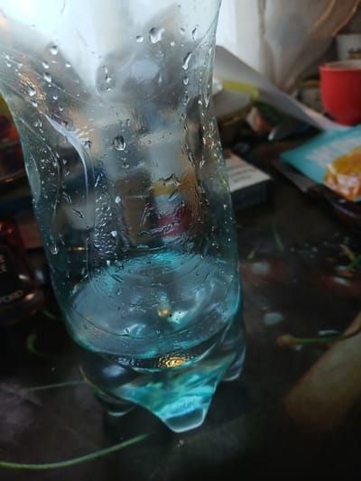 The image shows a close-up of a clear plastic bottle filled with a blue liquid. The bottle is covered in condensation. The background is blurred and appears to be an indoor setting, with objects such as a red cup, a pack of cigarettes, and what looks like food. There is no visible indication of the specific location within Hlushky, Ukraine. The scene appears to be a still life arrangement.