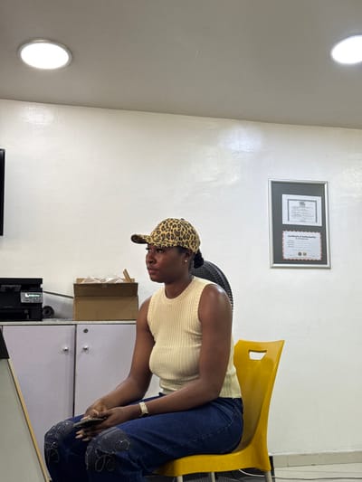 In an indoor setting within the city of Ajah, Nigeria, a young adult Black female is seated on a bright yellow plastic chair. She is oriented to the left of the frame, wearing a leopard-print baseball cap, a light cream ribbed sleeveless top, and dark blue jeans with black appliqué designs on the knees. A gold-toned watch adorns her left wrist, and she holds a dark-colored smartphone in her hands, which rest in her lap. Her gaze is directed towards the left. The background features a plain white wall and a light-colored ceiling with two illuminated circular recessed lights. To the left, a dark flat-screen monitor is mounted on the wall above a light gray cabinet holding a dark printer and a brown cardboard box containing items. A portion of a black standing fan is visible behind the subject's head. To the right, two framed documents, including one labeled "Certificate of Authorization" and another with "NBL," hang on the wall.