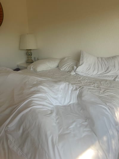 A close-up, slightly tilted shot captures a mostly white scene. A bed dominates the frame, covered in rumpled white bedding, including pillows and a duvet or comforter. To the left, a wall with a lamp is visible, with the lamp consisting of a white shade and a decorative base with blue and green accents. A small portion of a circular object is also visible at the very bottom left, hinting at possibly furniture.