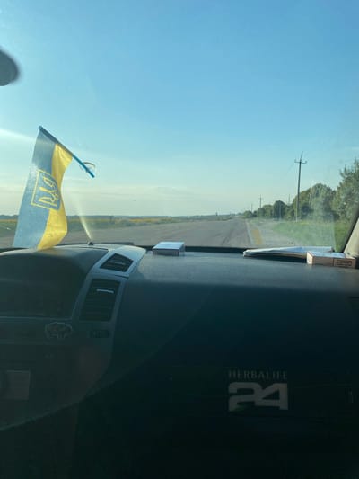 A first-person perspective shot from inside a car, looking through the windshield at a rural road. The dashboard is visible in the foreground, with a Ukrainian flag attached to the windshield on the left. Two packs of cigarettes and a folded piece of paper are on the dashboard. The road stretches into the distance, lined with trees and power lines. The sky is clear and blue. A Herbalife 24 logo is visible on the lower right of the dashboard. The scene is set in the city of Maiske, Ukraine, on a sunny day.