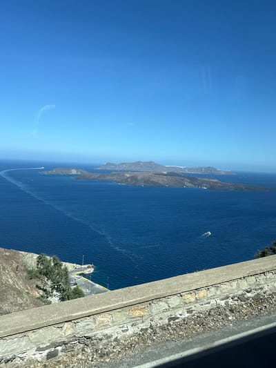 A high-angle shot captures a vast expanse of deep blue Aegean Sea under a clear, bright sky. In the foreground, a rough, stone-and-concrete retaining wall runs horizontally across the bottom of the frame, with a paved road visible below it. The wall's upper edge is lined with weathered concrete, and beneath it, a jumble of rocks and gravel is visible.

Beyond the wall, the sea dominates the scene. Several islands, appearing rugged and mountainous, are scattered across the water, their dark, rocky surfaces contrasting with the bright blue sea and sky. Some of these islands are lined with white buildings, suggesting a settlement. White wakes trail behind a small boat moving across the sea. In the mid-ground, a quay or harbor is visible, with a white ferry or tour boat docked alongside. A few other smaller boats are also present. Trees and sparse vegetation grow on the steep, rocky slopes leading down to the water.

The scene is likely from the caldera of Santorini, a volcanic island in Greece, possibly from a vantage point in Thira Municipal Unit. The distinctive geological formations and the presence of the caldera suggest this location.