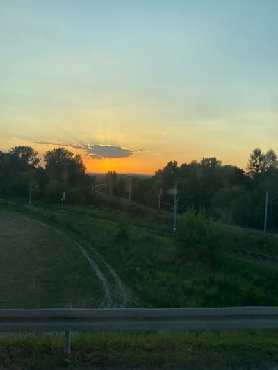 The image shows a landscape scene at dusk with a railway track curving through lush green vegetation and trees. The sky is a gradient of soft blues and yellows, with the sun partially obscured by a cloud, casting rays of light. A metal guardrail is visible in the foreground. The location is identified as Podłęże, Poland. There are no people or animals visible in the image.