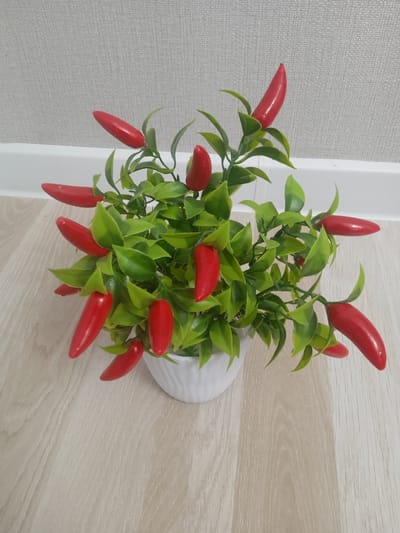 A close-up, high-angle shot shows a potted artificial plant with bright red chili peppers and green leaves. The plant is in a white ceramic pot with a textured, ribbed surface. The pot sits on a light-colored wooden floor with a visible wood grain pattern. In the background, a wall with a light gray, textured wallpaper is visible, with a white baseboard separating the wall from the floor. The lighting appears to be indoor, with no specific time of day or weather cues. The scene is static, with no activity taking place. There is no visible text in the image.
