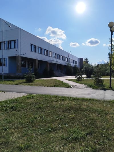 The image depicts a sunny day scene in Roshchinsky, Russia, focusing on the exterior of a modern, light-colored administrative building. The building features a long facade with numerous windows, a light blue lower section, and a prominent sloping roofline. A street lamp stands to the left of the building, and another one is visible in the distance to the right. The foreground consists of a paved walkway, a patch of green grass, and a small evergreen tree. The sky is a bright blue with scattered white clouds, and the sun is a bright disc near the top center of the frame. The overall impression is one of an organized, well-maintained public space under clear weather conditions.