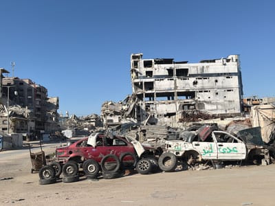 In Jabalia, Palestinian Territories, a scene of widespread destruction unfolds under a clear blue sky. The foreground is dominated by the remnants of vehicles and piled tires. A red sedan, missing its hood and parts of its interior, sits prominently, surrounded by several tires stacked and leaning against it. To its right, a white sedan, also heavily damaged and missing wheels, bears green Arabic graffiti on its side. Behind these vehicles, the landscape is characterized by collapsed buildings and rubble. A large, multi-story concrete structure, severely damaged with gaping holes where windows and floors once were, stands as a testament to the destruction. Other damaged buildings and debris are visible throughout the scene, indicating a significant impact from conflict or disaster. The ground is dusty and uneven, littered with small debris. In the background, glimpses of more damaged structures and tents suggest a populated area that has suffered extensive damage.