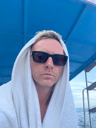 A man with fair skin and blond hair is wearing sunglasses and a white towel draped over his head. He appears to be on a boat, evidenced by the blue exterior of the vessel visible on the left, and a glimpse of the sea and a boarding ladder behind him. His expression is neutral.