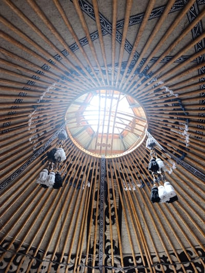 The image shows the interior of a yurt in Kegeti, Kyrgyzstan. The view is looking upwards towards the central opening, or "tunuk," of the yurt's roof. The structural elements of the yurt are visible, consisting of wooden poles radiating from the center to the circular walls (not fully shown). These poles are covered with a fabric liner, possibly felt or canvas, which is decorated with traditional geometric patterns in black and white. Several tassels, made of white, black, and grey yarn, hang from the wooden poles, adding a decorative touch. The "tunuk" itself is an opening to the sky, with visible wooden supports and some of the yurt's outer covering. Sunlight streams through the opening, illuminating the interior and casting light onto the structural elements. The overall impression is one of traditional nomadic architecture and decor.