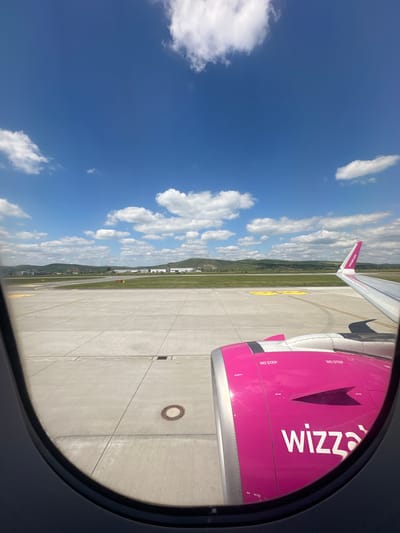A view from an airplane window on a sunny day, showcasing a Wizz Air plane on the runway, presumably at an airport near Ungheni, Romania. The aircraft engine is visible, emblazoned with the "Wizz" logo in white on a pink background. The plane's wingtip and tail fin are also visible. The runway is made of concrete with yellow lines marking the taxiways. Beyond the runway, there is a green field and a small mountain range. The sky is a bright blue with scattered cumulus clouds. The presence of the Wizz Air plane and the landscape of hills and a small city at the end of the runway suggest the location is likely in or near Ungheni, Romania.