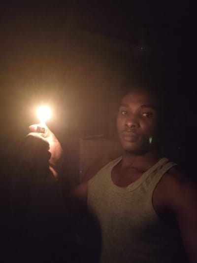 A young man with dark skin and short dark hair is illuminated by a lighter flame he is holding in his right hand. He is wearing a light green tank top. The background is dark, with some blurred objects that suggest an indoor environment. The man is looking towards the camera. The image appears to have been taken at night. The location in Orodo, Nigeria is unknown.