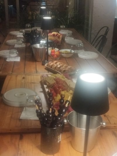 A long wooden table set for a meal displays plates, glasses, and several food arrangements. Skewered grapes in a mug, a charcuterie board with tomatoes and cheese, and bowls containing tomatoes and olives are visible. Mini sausage rolls are also placed along the table. Small black lamps provide lighting along the table. Chairs are present around the table, suggesting a ready-to-eat setup.