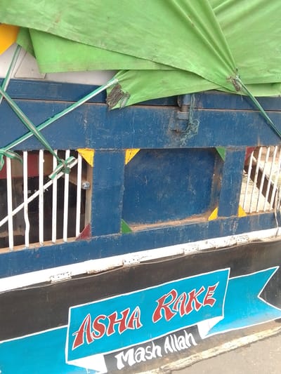 The image shows the side of a blue truck with a green tarp covering part of its load. The truck has white vertical bars on its side, forming a grate. The lower part of the truck has black and blue painted sections with text. The text in red and white says "ASHA RAKE" on a blue banner. Below this, in white cursive font, is the text "Mash Allah". The truck appears to be parked on a street, with a worn asphalt surface visible in the foreground. The scene suggests a commercial vehicle, possibly used for transport. The location is identified as Gulum, Nigeria.