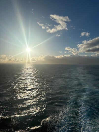 This image depicts a marine environment viewed from a moving vessel, likely off the coast of San Sebastián de La Gomera, Spain, given the faint landmass on the distant horizon. The scene is dominated by the ocean and sky. A brilliant, starburst-effect sun is positioned low in the left quadrant of the sky, reflecting intensely as a shimmering path across the dark blue and teal ocean surface, extending towards the foreground. The sky above is a clear blue with scattered white cumulus clouds, particularly along the horizon and in the upper right. A distinct, frothy white wake from the vessel from which the image was taken occupies the lower right portion of the frame, indicating active movement. No individuals or specific objects beyond the natural elements and the vessel's wake are visible. The overall impression is an open seascape under bright daylight conditions.