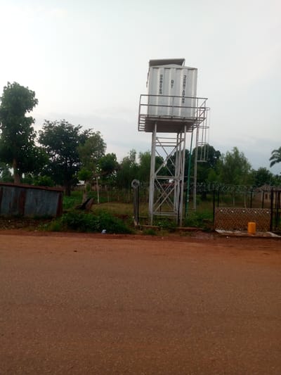 In Kaura, Nigeria, a tall, white water tank, inscribed with "DONATED BY JERRY ADAMS," stands elevated on a metal scaffold. Below the tank, a faucet system with tiled backing is visible, and a yellow container sits nearby. The scene is bordered by a fence topped with barbed wire. Green trees and a corrugated metal structure are present in the background. The ground in the foreground is an unpaved, reddish-brown road.
