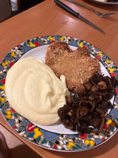 A close-up, overhead shot captures a meal on a wooden table. The plate, adorned with a colorful, patterned rim, holds a serving of creamy mashed potatoes, a breaded cutlet, and a pile of caramelized mushrooms and onions. A knife and fork rest on the table to the right of the plate. The setting appears to be a home environment in Skawina, Poland.