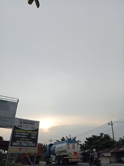 The image depicts a roadside scene in Kubang Jaya, Indonesia, under a hazy, overcast sky with the sun partially visible through the clouds, suggesting it is late afternoon or early morning. In the foreground, a large billboard displays "BAJA RINGAN INDONESIA WIJATRUSS" and "LOWONGAN PEKERJAAN" (Job Vacancy) with listings for Sales Marketing, Admin Logistik, Helper, and Driver, along with contact information. Another partially visible billboard to the left appears to be related to an educational institution, possibly mentioning "PESANTREN" (Islamic boarding school) and "RIAU".

To the right of the billboards, a Pertamina fuel tanker truck is parked on the road. The truck has the Pertamina logo and is labeled with information such as "PREMIUM", "SUPER", and a safety warning with the number "35". A motorcycle carrying two individuals, one wearing a white helmet, is traveling on the road in front of the fuel truck. In the background, there are trees and several buildings with corrugated metal roofs. The overall impression is of a commercial and transportation hub along a road in a semi-urban or rural area.