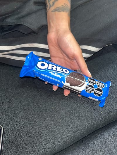 A human hand holds a blue, unopened package of Oreo Original cookies. The package has warning labels indicating excessive amounts of sugar, saturated fats, total fats, and calories. The background shows black fabric and black athletic pants with white stripes. The packaging indicates the product is from Argentina. The location of Llavallol cannot be determined from the image.