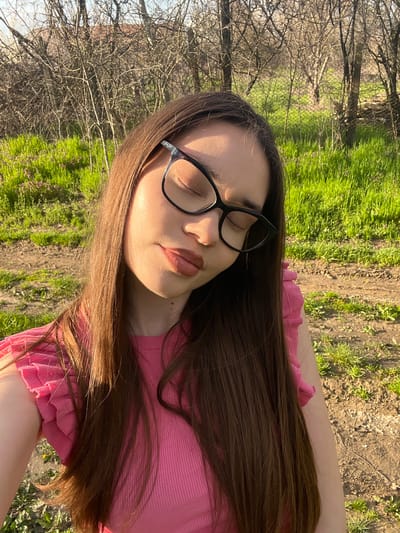 A young woman with long brown hair and wearing black-rimmed glasses lies on the grass outdoors. She has her eyes closed and is wearing a pink top. The background includes green grass, a dirt path, and bare trees, indicating a park or natural area. The image's orientation is vertical. It is taken at ground level. There is no identifiable information to confirm the location is Blaskovo, Bulgaria.