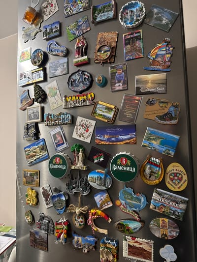 The image shows a collection of magnets attached to a metallic surface, likely a refrigerator door. The magnets are souvenirs from various locations and represent different landmarks, attractions, and cultural symbols. Some are labeled with names like "Barcelona", "Golden Sands", "Sunny Beach," "Plovdiv", "La Morra" and "Olympic Beach". Some feature texts in Bulgarian. Other magnets display images of landscapes, architecture, and people. There are bottle cap magnets with the word "Kameница". There is nothing in the image to suggest any association with the city of Gara Elin Pelin in Bulgaria.
