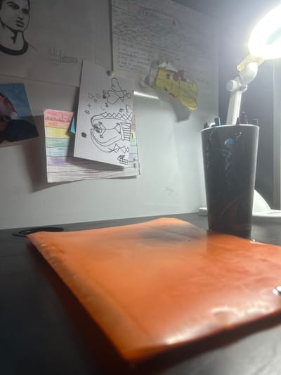 The image depicts a close-up, low-angle shot of a desk with various items related to a school or study environment. In the foreground, a shiny, orange folder or binder lies flat on the dark desk surface. To the right, a tall, dark cylindrical cup, possibly a pencil holder, contains writing utensils and is illuminated by a bright desk lamp positioned behind it. The lamp's light is diffused, casting a glow on the wall behind the desk.

The wall itself is adorned with several pieces of paper. On the left, a charcoal drawing of a person's head and shoulders is visible, with the name "Yora" written in cursive below it. Next to this, a photograph of a person is partially obscured, and below that, a small stack of colorful paper strips with the name "marcel c" written on one of them. To the right of the "Yora" drawing, a sheet of graph paper displays a black and white drawing of a cartoon character with large eyes and an open mouth, surrounded by hearts and other small shapes.

Further to the right on the wall, a sheet of paper contains handwritten text in French, with the heading "ENTRAINE-TOI". This document appears to be a list of instructions or goals, with bullet points detailing actions like "Je dois être gris", "Je dois avoir un mentalité muscler", "Je dois vivre [illisible]", and "Je dois m'entraîner tous les jours". There is also a small cutout image of Pikachu taped to the wall above this document. The overall impression is of a workspace that is personalized with artwork and study materials, possibly belonging to a student. The presence of French text and the name "Carrières-sous-Poissy" within the image metadata suggests this scene is located in that city in France.