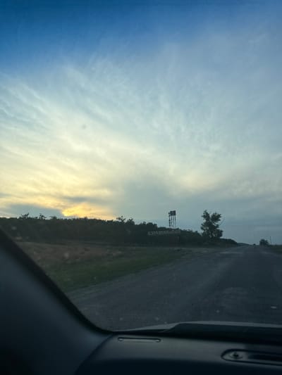 The image captures a rural roadside scene in Sadaclia, Moldova, likely during the late afternoon or early evening. The perspective is from inside a vehicle, looking out to the left. A grey asphalt road runs alongside a grassy verge bordered by trees. In the distance, a tall, metallic structure, possibly a radio tower or part of a communication array, stands against the sky. A solitary tree with sparse foliage is also visible near this structure.

The sky is a prominent feature, transitioning from a deep blue on the left to lighter shades of white and yellow towards the right, suggesting the presence of sunlight breaking through clouds or a sunset. Wispy clouds stretch across the sky, creating a dynamic and somewhat dramatic atmospheric effect.

On a wooden fence or structure along the roadside, there is visible text. It is printed vertically and appears to be in a dark color. While the text is partially obscured and angled, it reads "SALVETRANS" in a stylized font. The overall mood of the image is tranquil, evoking a sense of open space and the quiet beauty of the Moldovan countryside. The weather appears clear with a hint of warmth from the sun's low angle.