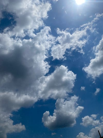 An extreme upward-facing shot captures a brilliant blue sky dotted with fluffy, white cumulus clouds. Sunlight streams in from the top right, creating a slight lens flare and casting a soft glow on the clouds. The clouds are varied in size and density, with some appearing dense and shadowed, hinting at their volume. The image is taken from Berangas Timur, Indonesia.