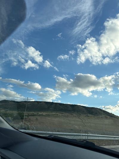 The image captures a landscape scene from within a vehicle in Krivi Dol, North Macedonia. A large, dark dashboard or car interior element dominates the foreground, partially obscuring a reflective surface, likely a windshield. Beyond this, a bright blue sky is filled with a variety of clouds, from wispy cirrus formations to more substantial cumulus clouds.

The middle ground features a hilly, arid landscape. The hills are covered in dry, light brown vegetation, with some darker green patches indicating sparse shrubbery. Distinctive erosion patterns are visible on the slopes. A metal guardrail runs horizontally across the scene, separating the foreground from the distant landscape. On the hillsides, there are streetlights. One prominent streetlight stands tall, positioned slightly to the left of the center.

The overall environment suggests a daytime scene, with the sun illuminating the landscape and sky. The view appears to be from a moving vehicle on a road, as indicated by the perspective and the elements in the foreground.