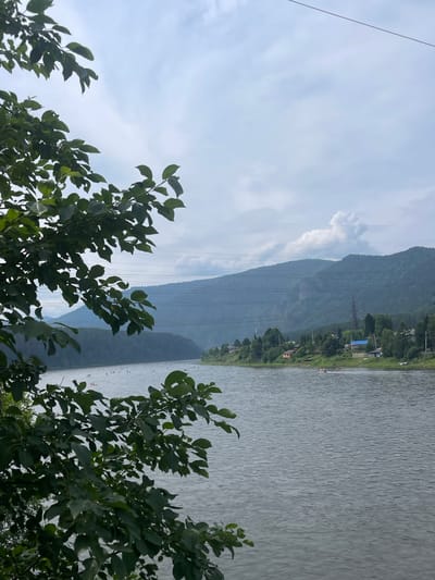 This image captures a serene view of the Mana River in Ust-Mana, Russia. In the foreground, lush green tree branches with broad leaves partially obscure the scene. The river itself is a wide expanse of calm, gray water, with visible ripples suggesting a gentle current.
Along the far bank of the river, a settlement is visible, characterized by a dense line of trees interspersed with scattered houses and buildings, some with blue roofs. Several power lines stretch across the mid-ground, supported by tall pylons. Beyond the settlement, rolling, tree-covered mountains rise in the background, fading into a hazy, overcast sky.
On the water, numerous small, light-colored objects, likely kayaks or paddleboards, are scattered across the river, indicating recreational activity. A few people are visible on the far bank near the settlement, some appearing to be resting or sunbathing. A small motorboat can be seen on the river. The overall atmosphere is one of tranquil natural beauty with signs of human habitation and leisure.