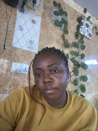 The image displays an adult individual, identified as female, with dark skin, braided hair (including some reddish-brown strands), a nose piercing on her left nostril, and an earring in her right ear. She is wearing a mustard yellow long-sleeved top and is looking directly at the camera.

The background is an indoor wall featuring a light brown and beige textured pattern, possibly wallpaper. Mounted on this wall are several items:
- In the upper left, a black electrical cable is plugged into a white wall socket, with another black cable hanging downwards.
- Below this, a white rectangular decorative plaque or tile displays a black grid pattern and illustrations of a coffee cup and a potted plant.
- In the lower left, a white electrical switch and multiple-socket unit is installed. A white charger is plugged into one socket, with a white cable extending from it.
- In the upper right, a circular, silvery decorative object, possibly woven metal, is adorned with artificial green vine leaves that trail downwards.
- To the right of the circular decoration, a rectangular decorative piece with a red and white patterned design is affixed.
- In the lower right, another white electrical socket is visible.

The electrical fittings are consistent with those found in Nigeria. The image does not provide any identifiable landmarks or specific features to ascertain its precise location within Abuja, Federal Capital Territory, Nigeria.
