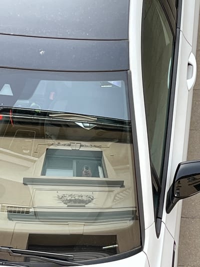 Overhead shot of the white roof and side windows of a car. The reflection in the side window shows a light-colored building with a distinctive window and decorative detailing. The location of the image is Foetz, Luxembourg.