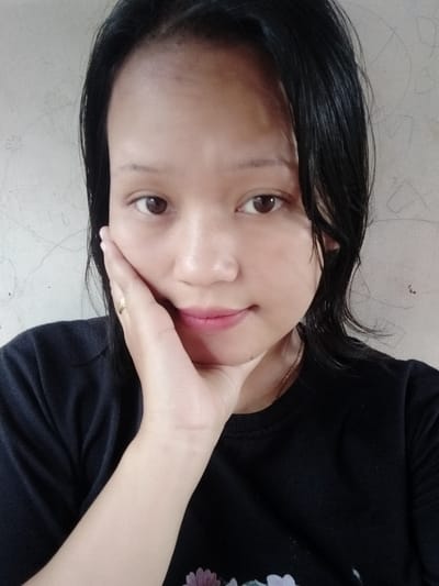 A young woman with dark hair poses indoors, her hand gently touching her cheek. Her expression is soft, and she wears a dark-colored t-shirt. A gold ring is visible on her finger. The background is a plain, light-colored wall. Her hair is short and appears to be wet. She wears light pink lip color, and her eyes are brown. A thin gold necklace is visible around her neck.