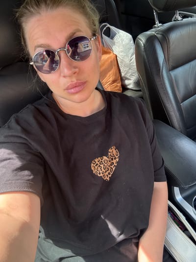 A medium shot captures a woman in a car, seated in the driver's seat, facing the camera. She is wearing a black t-shirt with a leopard print heart graphic on the left side of her chest and round, silver-rimmed sunglasses. Her blonde hair is pulled back. The interior of the car appears to be black leather. In the background, on the passenger side, are two plastic shopping bags, one orange and one white. The image was taken in the city of Valday, Russia.