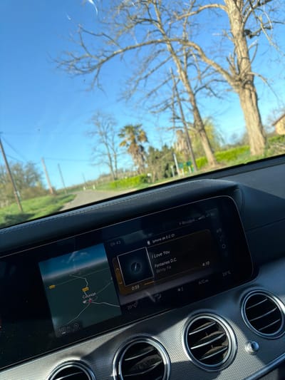 The scene is captured from inside a vehicle, looking forward over the dashboard. The car's infotainment screen displays a navigation map on the left, centered on the location "Cucasse," which is situated within the commune of Mauléon-d'Armagnac, France, specifically showing roads D223 and D114. To the right, the screen shows music playing, identified as "I Love You" by Fontaines D.C. from the album "Skinty Fia," connected via "Iphone de E.C (2)," with 0:55 elapsed of a 4:10 track. The time 09:43 is visible, along with temperature readings of 20°C and 23°C. Below the screen, multiple circular air vents are present. Through the windshield, a narrow, paved road extends straight ahead into a rural landscape. The roadside is lined with green grass and various trees, including mature deciduous trees with bare branches, a palm tree, and other trees with green foliage. Utility poles with overhead lines are visible along the road's edge, under a clear blue sky.