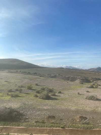 The image displays a vast and rugged natural landscape under a bright, clear blue sky, viewed in correct landscape orientation. The sky, occupying the upper half, features a vibrant blue gradient with subtle, wispy white clouds stretching across.
The lower half reveals an expansive, gently undulating terrain composed primarily of rocky and gravelly ground. Scattered across this textured surface are sparse patches of green vegetation, appearing as small shrubs or bushes. These plants cast distinct, elongated shadows, indicating strong, directional sunlight, characteristic of morning or late afternoon.
In the mid-distance and background, a chain of low-lying mountains or hills extends across the horizon, appearing hazy and lighter in color due to atmospheric perspective. A faint line, possibly a utility line with poles, can be observed running along the base of these distant formations.
To the far right of the foreground, a segment of a concrete barrier or low wall is visible, with faint indications of a fence structure nearby. A small, white, angular object is also present on the rocky ground in the mid-right.
No people or specific activities are depicted, lending the scene an undisturbed and tranquil atmosphere. The arid, rocky terrain with distant mountains is consistent with the provided location context of Jbilate الجبيلات, Morocco, showcasing its natural, undeveloped beauty.