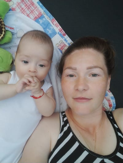 This is a selfie taken from above, showing a woman and a baby. The woman is in the foreground, looking at the camera with a slight smile. She has light skin with freckles and her dark hair is pulled back. She is wearing a black and white striped tank top and a delicate gold necklace. To her left is a baby, lying on its back and looking towards the camera. The baby is wearing a white onesie and has its hands near its mouth. A red bracelet is visible on the baby's wrist.
They are lying on a patterned blanket, which has red, white, and blue geometric designs and some cartoon characters. To the left of the baby, two green plush turtle toys are visible. The background appears to be a dark, textured surface, possibly a bed or mat. The lighting suggests it is daytime, and the overall mood is relaxed and intimate. The location context is Fălești, Moldova, but there are no specific visual cues in the image to confirm this. There is a visible logo on the white blanket that says "Bugel".