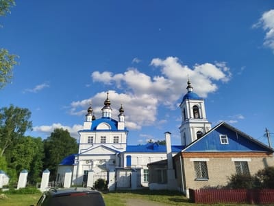 The image shows a church in Мамонтово, Russia, under a blue sky with scattered clouds. The church is predominantly white with blue roofs and domes topped with golden crosses. A bell tower rises on the right side of the main structure. A brick building with a blue roof is adjacent to the church on the right. A red fence surrounds the brick building. A black vehicle is partially visible in the foreground. Trees and greenery surround the structures.