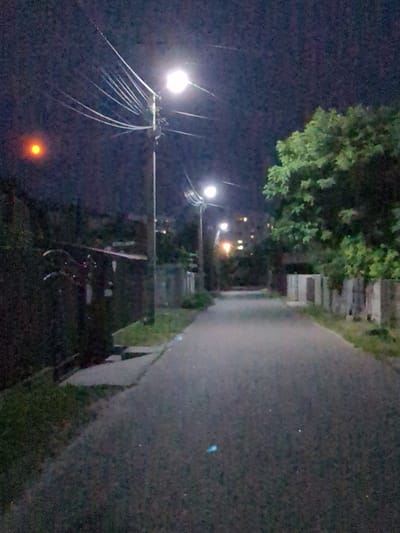 The image shows a deserted, dimly lit street at night, likely in the city of Boryspil, Ukraine, as indicated by the provided context. A prominent street lamp illuminates the asphalt road, casting long shadows. To the left, a dark fence and some greenery line the street, with a faint orange circular light visible in the sky, possibly the moon or a distant light source. On the right side of the road, there is a stone wall and a dense tree with green foliage. Further down the road, several other street lights and building lights are visible, suggesting the presence of a residential or urban area. The overall atmosphere is quiet and somewhat eerie, typical of a nocturnal urban scene.