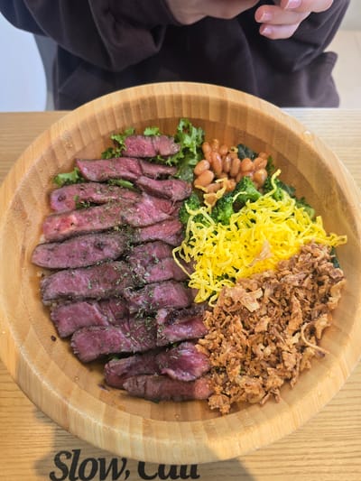 This media file is an overhead shot of a wooden bowl filled with food. The bowl contains sliced steak, shredded yellow egg, broccoli florets, baked beans, and crispy fried onions. The steak is seasoned with herbs and appears to be cooked medium-rare. The food is arranged in sections within the bowl, creating a colorful and appetizing presentation.

To the left of the bowl, a person's hand with painted fingernails is visible, suggesting that someone is about to eat or has just finished eating the meal. The hand is resting on a dark brown garment. The setting appears to be indoors, with a wooden tabletop and a light-colored surface visible below the bowl.

On the right side of the wooden bowl, black text is visible. It reads "Slow, Cuu". The lighting suggests it could be daytime, possibly with natural light filtering in, as there are some bright highlights on the bowl and food. The overall mood is casual and suggests a mealtime setting. The location context provided is Gumi-si, South Korea.